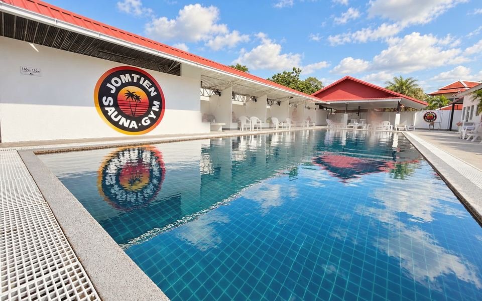 The outdoor pool at Jomtien Sauna & Gym on a sunny day, with the round Jomtien Sauna Gym logo on the white wall and red-roofed cabanas along the deck