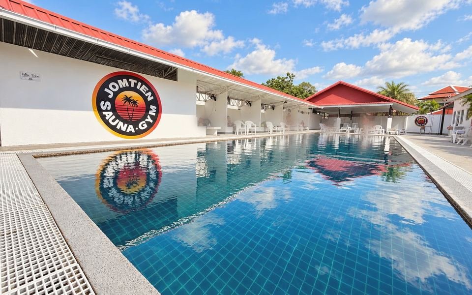 Открытый бассейн Jomtien Sauna & Gym в солнечный день: круглый логотип на белой стене и беседки под красной крышей вдоль бортика